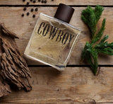 Glass bottle of Cowboy cologne on rustic wood with green pine branches and natural elements.