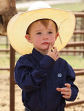 Cinch toddler boy wearing a royal blue western shirt and straw cowboy hat, looking thoughtful on a ranch.