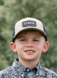 Cinch Boys Trucker Cap worn by a young boy, featuring a beige front and navy mesh backing, outdoors.