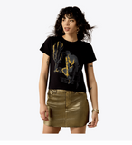 Model wearing Ariat Women's Wild West Short Sleeve Graphic T-Shirt with bold graphic, paired with a shiny gold skirt.